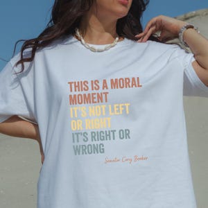 Op de afbeelding: Wit T-shirt met de tekst "THIS IS A MORAL MOMENT IT'S NOT LEFT OR RIGHT IT'S RIGHT OR WRONG" in verschillende kleuren. De tekst wordt toegeschreven aan senator Cory Booker. Het shirt is een casual kledingstuk.