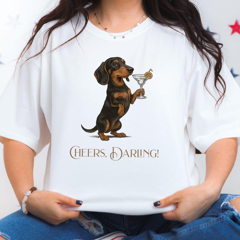 May include: White t-shirt featuring a dachshund dog illustration holding a martini glass. The dog is black and brown. The text "Cheers, Darling!" is printed in brown script below the dog.