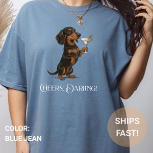 May include: Blue jean-colored t-shirt featuring a dachshund dog illustration holding a martini. The dog is brown and black. The text "Cheers, Darling!" is printed below the dog. The shirt is a relaxed fit.