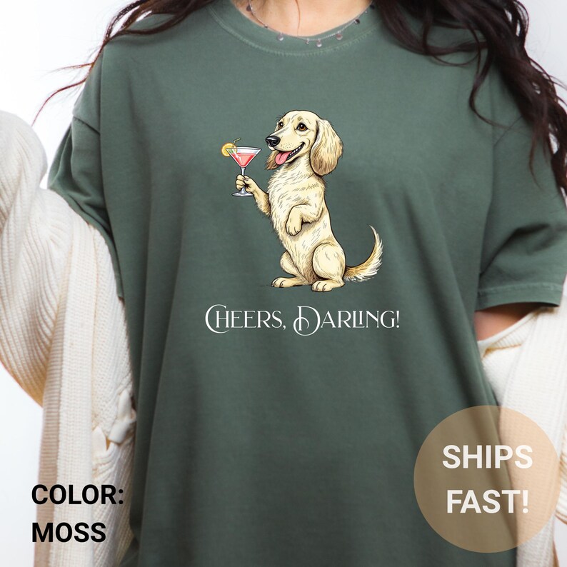 May include: Moss green t-shirt featuring a cartoon dachshund holding a cocktail. The dog is light tan with a pink drink in a martini glass. The text "Cheers, Darling!" is below the dog.
