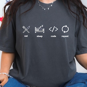 May include: Dark grey t-shirt featuring white graphic icons and text. The icons depict a fork and knife, a bed with a sleeping figure, code brackets, and a circular arrow. The text reads "eat, sleep, code, repeat."