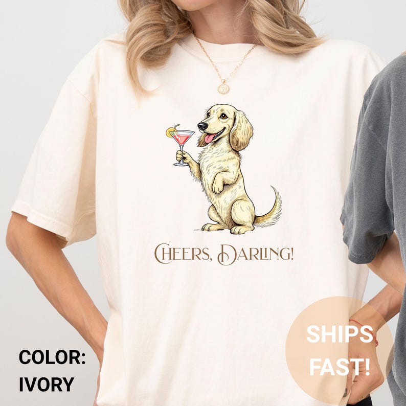 May include: Ivory-colored t-shirt featuring a cartoon dog holding a cocktail. The dog is light brown with floppy ears and a smiling expression. The text "Cheers, Darling!" is printed below the dog.