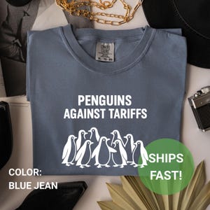 May include: A blue jean colored t-shirt with the text "PENGUINS AGAINST TARIFFS" in white. Below the text is a graphic of several penguins. The shirt also has a tag that says "COMFORT COLORS".