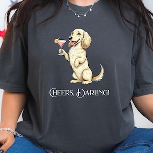 May include: Dark gray t-shirt featuring a cartoon dog holding a pink cocktail with a lemon garnish. The dog is standing upright and smiling. The text "Cheers, Darling!" is printed below the dog in white.