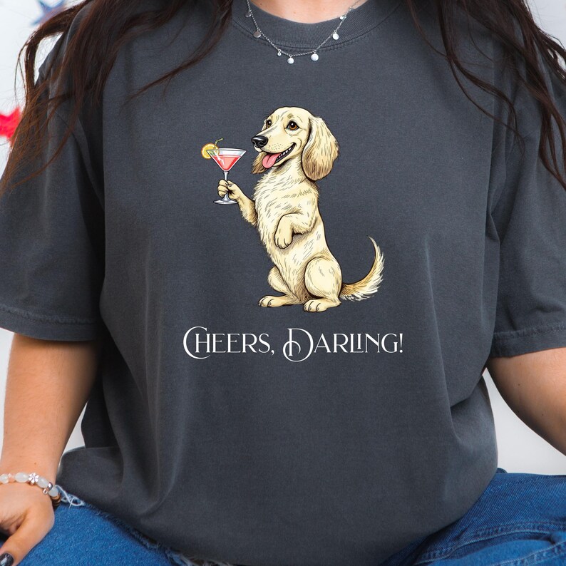 May include: Dark gray t-shirt featuring a cartoon dog holding a pink cocktail with a lemon garnish. The dog is standing upright and smiling. The text "Cheers, Darling!" is printed below the dog in white.