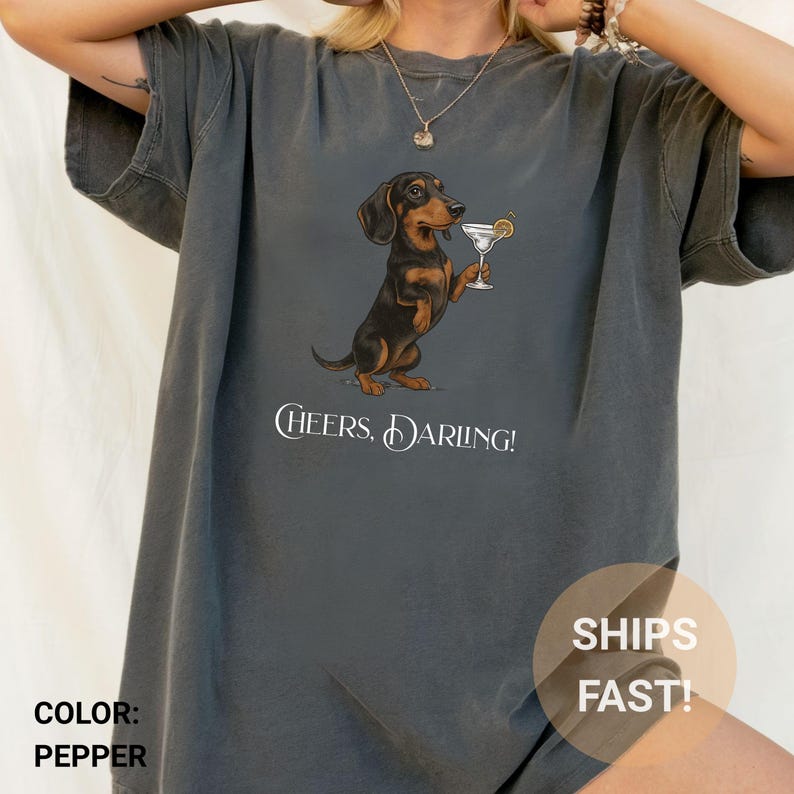 May include: Pepper-colored t-shirt featuring a dachshund dog holding a martini glass with a lemon garnish. The dog is black and brown. The text "Cheers, Darling!" is printed below the dog.