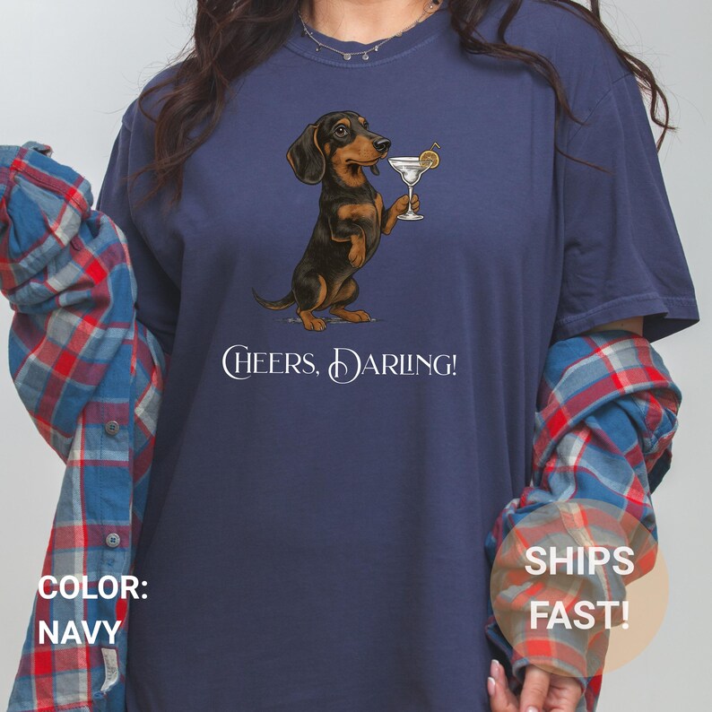 May include: Navy blue t-shirt featuring a dachshund dog holding a martini glass. The dog is brown and black. The text "Cheers, Darling!" is printed below the dog. The shirt has long sleeves.