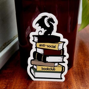 Puede incluir: Un tarro de vela rojo oscuro con tapa dorada. Una pegatina en el tarro presenta un dragón negro sobre una pila de libros. Los libros tienen las palabras "anti-social" y "bookclub". Una planta verde está en el fondo.