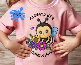 May include: A light pink t-shirt featuring a cartoon bee with the text "ALWAYS BEE GROWING". The bee is surrounded by colorful flowers. The shirt has short sleeves and is being held by a person.