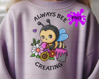 May include: A light purple sweatshirt featuring a cartoon bee with the text "ALWAYS BEE CREATING". The bee is holding a paintbrush and surrounded by flowers and a paint bucket. A purple heart and a purple PNG graphic are also present.