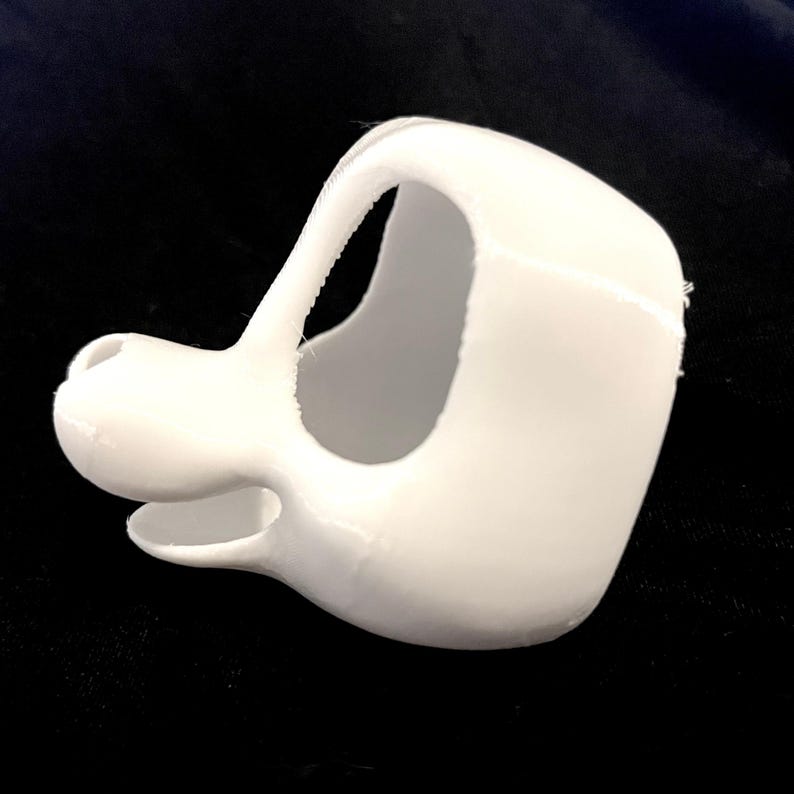 May include: A white, 3D-printed object resembling a stylized dog muzzle or mask. The object has a rounded snout, open mouth, and a curved handle or strap. The design is smooth with visible layer lines from the printing process.