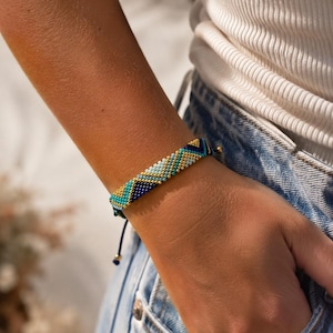 May include: A colourful beaded bracelet featuring a geometric design in teal, gold, navy, and light blue. The bracelet is worn on a wrist, with a navy blue adjustable cord and gold-coloured bead accents. A fashion accessory.