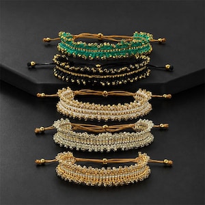 Könnte beinhalten: Fünf Armbänder mit Perlen in Grün, Schwarz, Weiß und Gold. Jedes Armband hat ein gewebtes Design mit kleinen Perlen und verstellbaren Kordelverschlüssen. Die Armbänder werden auf einer dunklen Oberfläche präsentiert.