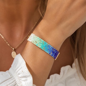 May include: A beaded bracelet with a gradient design of white, teal, and blue hues. The bracelet is rectangular and has gold-colored metal ends. A gold necklace is visible in the background.