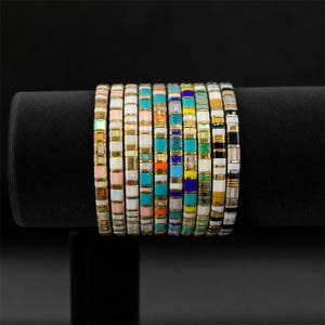 Op de afbeelding: Een verzameling kleurrijke tegelarmbanden die worden tentoongesteld op een zwarte fluwelen standaard. De armbanden hebben een verscheidenheid aan kleuren, waaronder roze, wit, goud, turquoise, oranje, blauw, geel en zwart. Elke armband is gemaakt van kleine, rechthoekige tegels.