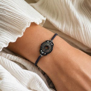 May include: A dark blue bracelet with an oval, beaded centerpiece. The centerpiece features a radial design with a central opening, surrounded by black beads and silver accents. The bracelet is worn on a person's arm, with a white fabric in the background.