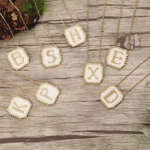 Può includere: Collane con catena color oro con ciondoli di perline bianche, ognuno con una lettera diversa in oro. I ciondoli sono di forma quadrata con un bordo dorato. Le collane sono disposte su una superficie di legno.