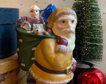 May include: A vintage ceramic Santa Claus figurine with a yellow and brown painted suit, carrying a green sack filled with toys, including a doll and a dog.