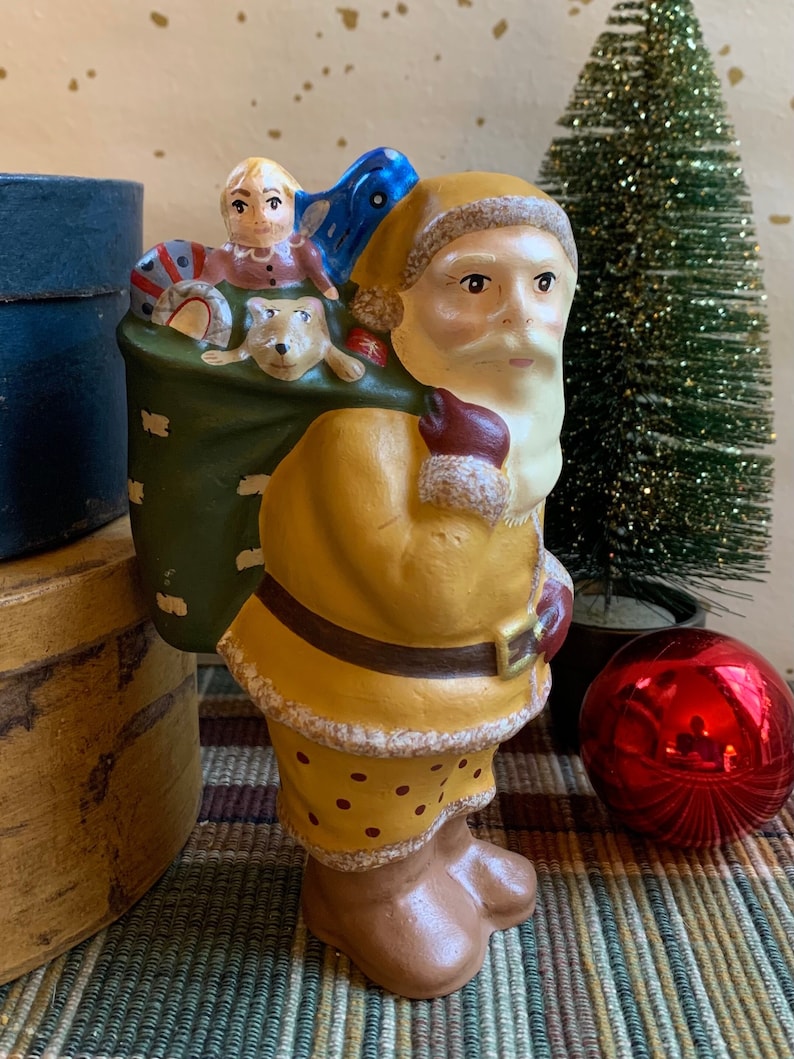 Pode incluir: Uma figura vintage de cer&acirc;mica do Papai Noel com um terno pintado de amarelo e marrom, carregando um saco verde cheio de brinquedos, incluindo uma boneca e um cachorro.