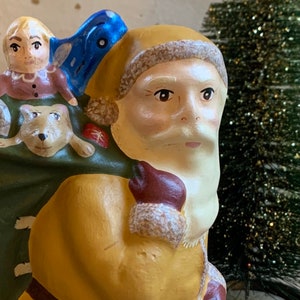Pode incluir: Uma figura de cer&acirc;mica vintage do Papai Noel com barba branca e casaco amarelo. Ele est&aacute; carregando uma bolsa verde com uma menina pequena e um cachorro dentro. A figura &eacute; decorada com detalhes azuis e vermelhos.