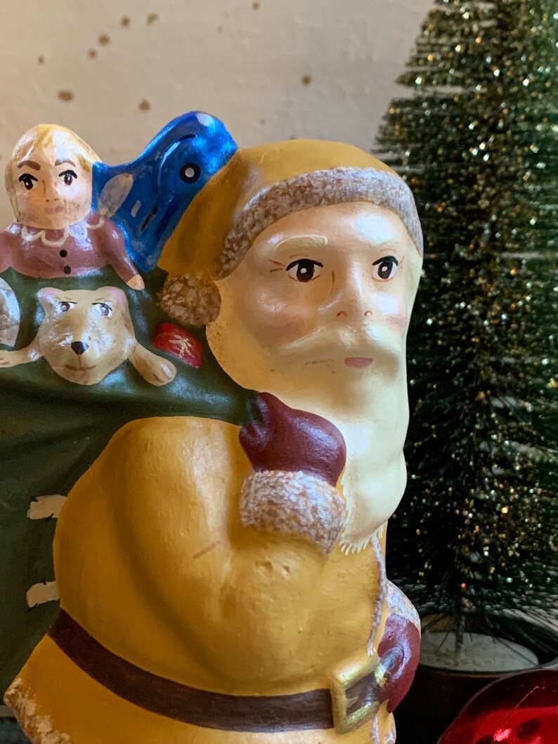 Pode incluir: Uma figura de cer&acirc;mica vintage do Papai Noel com barba branca e casaco amarelo. Ele est&aacute; carregando uma bolsa verde com uma menina pequena e um cachorro dentro. A figura &eacute; decorada com detalhes azuis e vermelhos.