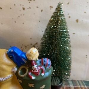 Pode incluir: Uma figurinha de camelo de cer&acirc;mica com um saco verde nas costas. O saco est&aacute; cheio de uma boneca de palha&ccedil;o, uma bola listrada azul e branca e um chap&eacute;u amarelo e branco. O camelo &eacute; pintado de amarelo com detalhes marrons.
