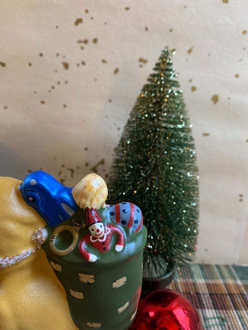 Pode incluir: Uma figurinha de camelo de cer&acirc;mica com um saco verde nas costas. O saco est&aacute; cheio de uma boneca de palha&ccedil;o, uma bola listrada azul e branca e um chap&eacute;u amarelo e branco. O camelo &eacute; pintado de amarelo com detalhes marrons.
