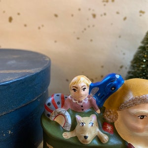 Pode incluir: Uma figura de cer&acirc;mica do Papai Noel com um saco verde cheio de presentes. Uma crian&ccedil;a pequena e um cachorro est&atilde;o sentados no saco. A crian&ccedil;a est&aacute; vestindo um vestido rosa e o cachorro &eacute; marrom e branco.