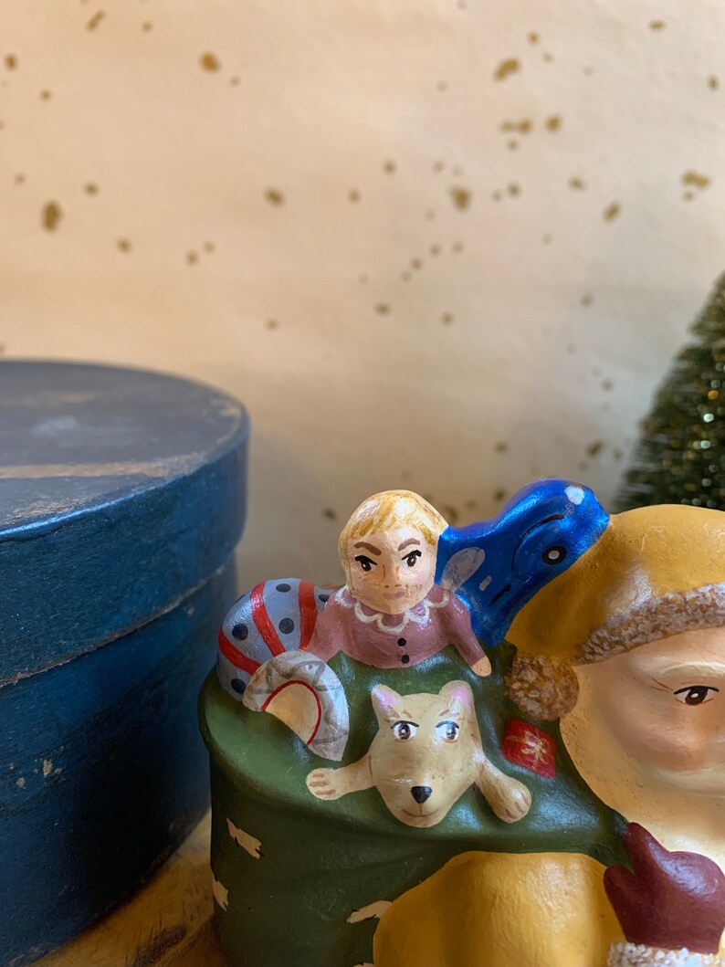 Pode incluir: Uma figura de cer&acirc;mica do Papai Noel com um saco verde cheio de presentes. Uma crian&ccedil;a pequena e um cachorro est&atilde;o sentados no saco. A crian&ccedil;a est&aacute; vestindo um vestido rosa e o cachorro &eacute; marrom e branco.