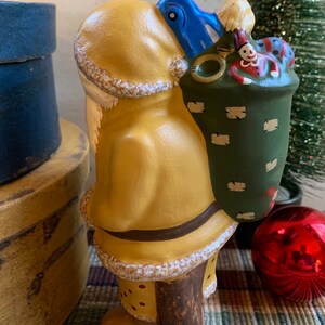 Pode incluir: Uma figura de cer&acirc;mica de Papai Noel pintada de amarelo com um chap&eacute;u azul e um saco verde cheio de brinquedos. A figura est&aacute; em p&eacute; sobre uma base de madeira e tem uma barba branca.
