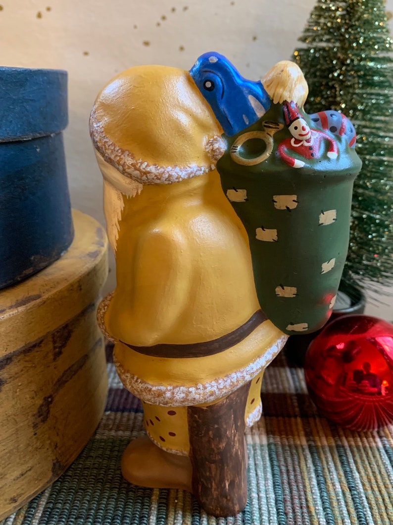 Pode incluir: Uma figura de cer&acirc;mica de Papai Noel pintada de amarelo com um chap&eacute;u azul e um saco verde cheio de brinquedos. A figura est&aacute; em p&eacute; sobre uma base de madeira e tem uma barba branca.