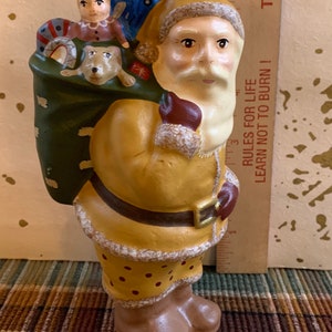 Pode incluir: Uma figura de cer&acirc;mica do Papai Noel carregando um saco cheio de presentes. O Papai Noel est&aacute; vestindo um terno amarelo e marrom com barba branca e chap&eacute;u vermelho. O saco &eacute; verde e tem uma fita azul. A figura tem aproximadamente 15 cm de altura.