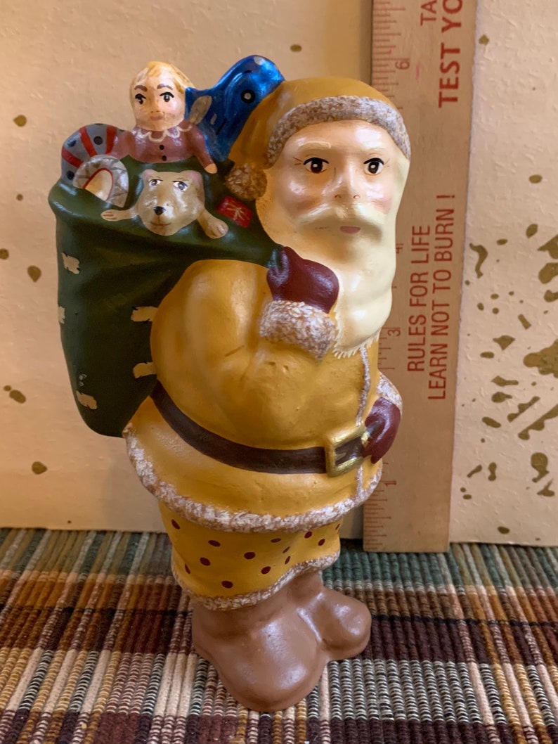 Pode incluir: Uma figura de cer&acirc;mica do Papai Noel carregando um saco cheio de presentes. O Papai Noel est&aacute; vestindo um terno amarelo e marrom com barba branca e chap&eacute;u vermelho. O saco &eacute; verde e tem uma fita azul. A figura tem aproximadamente 15 cm de altura.
