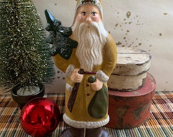May include: A vintage-style Santa Claus figurine with a long white beard, holding a green tree and a sack. The figurine is dressed in a mustard-colored coat and brown boots. A red Christmas ornament and a small green tree are in the background.