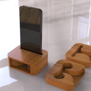 May include: A wooden phone stand with a slot to hold a phone upright. Two wooden coasters with the letters "A" and "B" are also present. The items are made of light brown wood and sit on a white surface.