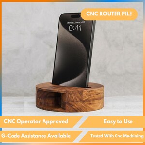 Puede incluir: Un soporte de teléfono de madera con una ranura para un teléfono inteligente. El teléfono muestra la hora y la fecha. El soporte es de madera marrón claro y tiene el texto "CNC ROUTER FILE". El texto adicional incluye "CNC Operator Approved", "Easy to Use" y "G-Code Assistance Available".
