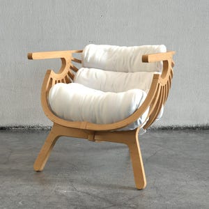 May include: A contemporary wooden armchair with a curved back and armrests. The chair has a light wood frame and two white, rectangular cushions for the seat and back. The chair is set against a neutral grey wall.
