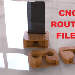 May include: Wooden phone stand with a slot for a phone, and two additional wooden pieces. The stand is made of light brown wood. The text "CNC ROUTER FILES" is in red.