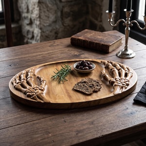 K&ouml;nnte beinhalten: Ein ovales Serviertablett aus Holz mit geschnitzten Skeletth&auml;nden an beiden Enden. Das Tablett enth&auml;lt eine kleine Keramikschale mit dunklen Oliven, einen Zweig frischen gr&uuml;nen Rosmarin und drei Scheiben dunkelbraunes K&ouml;rnerbrot auf der Oberfl&auml;che.