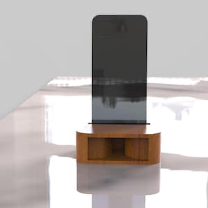 May include: A modern, minimalist phone stand. The stand features a dark gray rectangular screen supported by a rounded, light brown wooden base with a rectangular cutout. The stand is on a white surface.