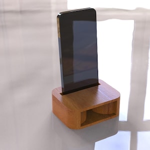 May include: A wooden phone stand with a slot to hold a smartphone upright. The stand is made of light brown wood and has a rectangular base with a curved top. The phone is black and has a rectangular shape.
