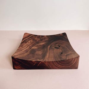 K&ouml;nnte beinhalten: Quadratisches Holztablett mit geschwungenem Rand aus dunkelbraunem Holz. Die Oberfl&auml;che zeigt eine ausgepr&auml;gte Maserung. Schlichtes, elegantes Design, geeignet zum Servieren oder Pr&auml;sentieren, ca. 25 cm x 25 cm.