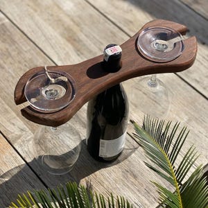 May include: A dark wooden wine glass and bottle holder with two circular cutouts for glasses. The holder sits atop a dark wine bottle with a cork. Two wine glasses are suspended from the holder. The background is a wooden surface.