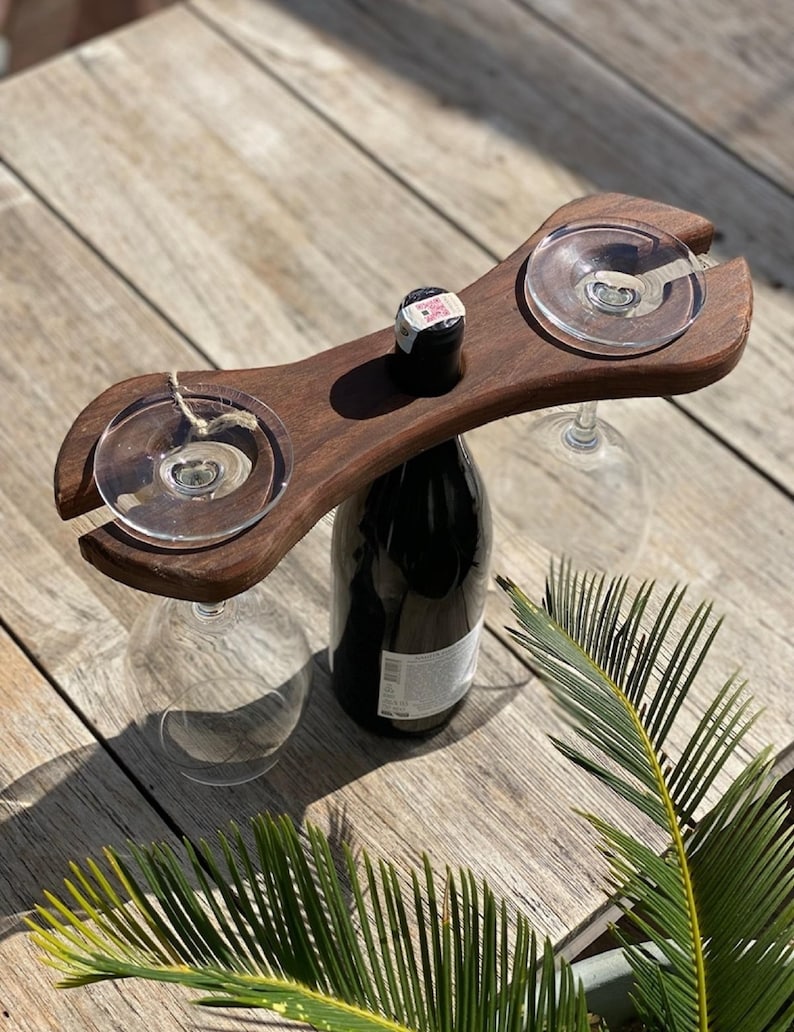 May include: A dark wooden wine glass and bottle holder with two circular cutouts for glasses. The holder sits atop a dark wine bottle with a cork. Two wine glasses are suspended from the holder. The background is a wooden surface.