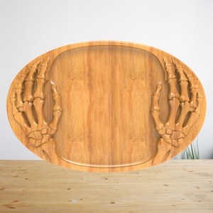 K&ouml;nnte beinhalten: Ein ovales Serviertablett aus Holz mit einem geschnitzten Reliefdesign aus zwei Skeletth&auml;nden auf der linken und rechten Seite. Das Tablett hat eine glatte, hellbraune Holzmaserung mit einer vertieften Mitte zum Abstellen.