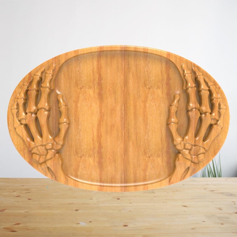 K&ouml;nnte beinhalten: Ein ovales Serviertablett aus Holz mit einem geschnitzten Reliefdesign aus zwei Skeletth&auml;nden auf der linken und rechten Seite. Das Tablett hat eine glatte, hellbraune Holzmaserung mit einer vertieften Mitte zum Abstellen.