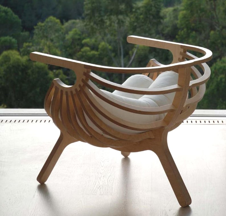 May include: A contemporary wooden chair with a distinctive design. The chair has a curved back and armrests, with vertical wooden slats creating the back and sides. The seat is a white cushion. The chair is made of light-coloured wood and has three legs.