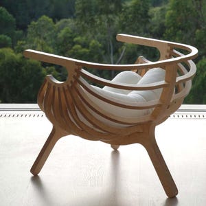 May include: A contemporary wooden chair with a distinctive design. The chair has a curved back and armrests, with vertical wooden slats creating the back and sides. The seat is a white cushion. The chair is made of light-coloured wood and has three legs.