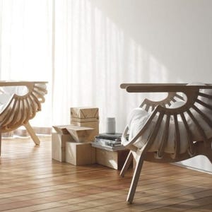 May include: Two chairs with a distinctive fan-shaped design, crafted from light-coloured wood. Each chair has a curved back and a cushioned seat. They are positioned on a wooden floor, with a white curtain visible in the background.