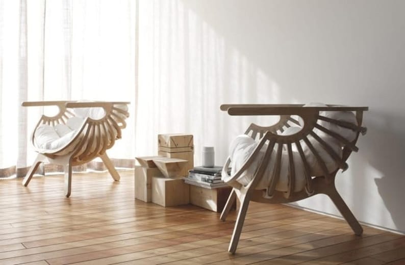 May include: Two chairs with a distinctive fan-shaped design, crafted from light-coloured wood. Each chair has a curved back and a cushioned seat. They are positioned on a wooden floor, with a white curtain visible in the background.